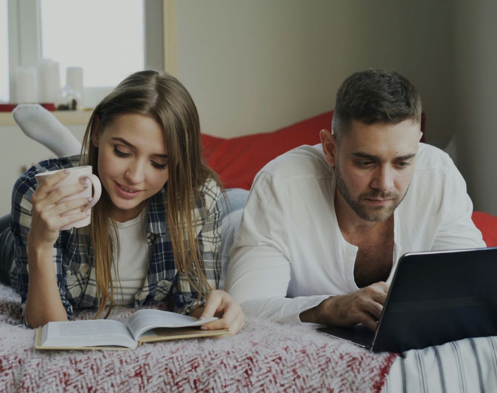 Un jeune homme et une jeune femme couchés côte à côte, à plat ventre sur leur lit. L'homme est devant son ordinateur, la femme lit.