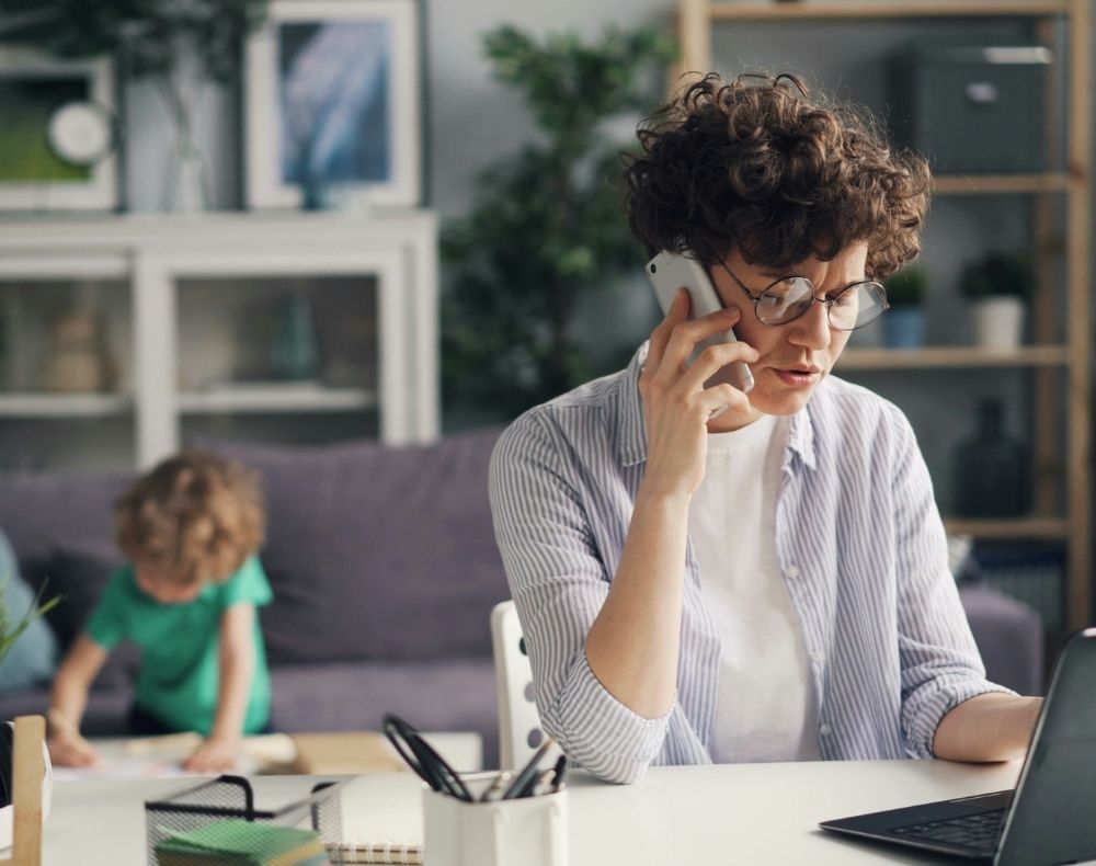 Femme devant un ordinateur qui est au téléphone. Un enfant joue en arrière-plan